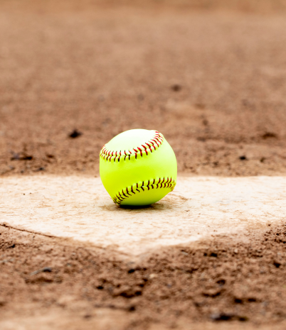 A yellow softball sits on home plate with the red dirt infield in the background.