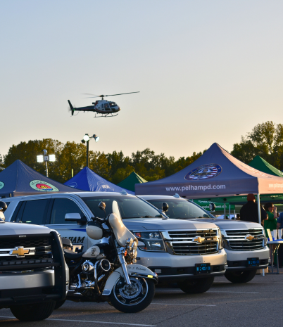 Rows of police vehicles, and tents are set up for National Night Out. A helicopter hovers overhead.